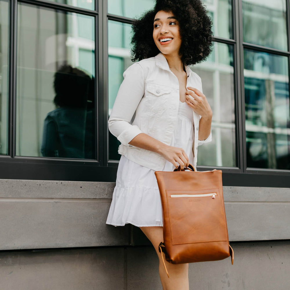 Smiling woman leaning against building holding a brown leather backpack